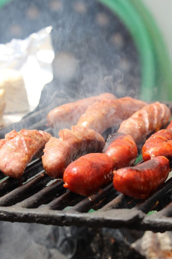 Closeup of a variety of juicy sausages grilling on an outdoor barbecue, surrounded by smoke.