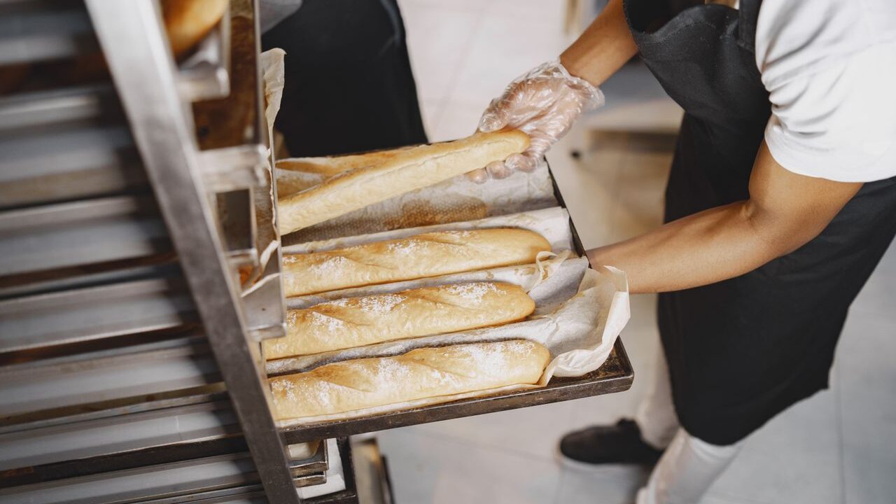 A baker arranges freshly baked baguettes on a rack, wearing gloves for hygiene.
